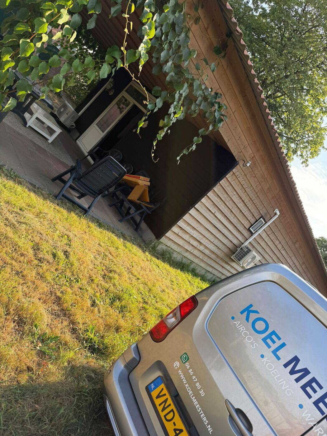 Buitenunit aan een houten bijgebouw in de tuin met de Koelmeesters-bus op de voorgrond